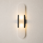 Vertical frosted wall sconce with black side brackets glowing warmly on a pale wall.