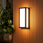 Glowing rectangular wall sconce on a vertical slat wall beside green indoor plant leaves.