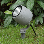 Gray outdoor spotlight with water droplets mounted on a stake in wet grass.