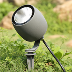 Gray outdoor spotlight on a ground stake among green grass and small plants.