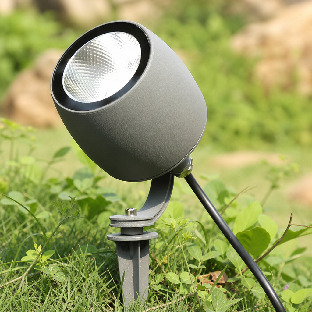 Gray outdoor spotlight on a ground stake among green grass and small plants.