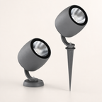 Two gray cylindrical outdoor spotlights with black rims and silver reflectors, one on a base and one on a ground spike.