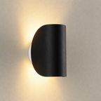 Black half-cylinder wall sconce casting warm light on a textured neutral wall.