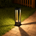 Black rectangular garden path light glowing on grass beside a brick walkway.