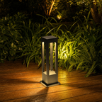 Black rectangular outdoor bollard light glowing warmly on a wooden deck beside green garden foliage.