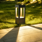 Black rectangular outdoor bollard lamp glowing on grass beside a paved path.