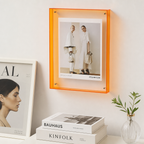 Orange acrylic wall frame displaying a Celine fashion poster above white books and a small plant.