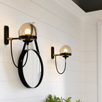 Two black-and-brass wall sconces with smoked glass globes and a round black mirror on a white paneled wall.