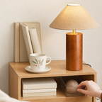 Wooden table lamp with beige shade on a shelf beside a mug, books, and a yarn box.