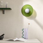 Green wall lamp above a lavender vase with wavy teal handle on a white tabletop.