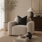 Cream boucle lounge chair with rounded arms and a dark charcoal pillow in a warm neutral living room.