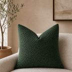 Dark green textured square throw pillow on a cream boucle chair in a neutral living room.