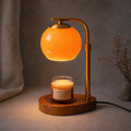 A brass candle warmer lamp with an orange glass shade illuminates a jar candle on a wooden base.