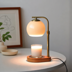A brass candle warmer lamp with an orange glass shade sits on a table, warming a white candle on its wooden base.