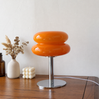 Glossy orange double-donut table lamp with chrome base on a wooden surface against a pale wall.