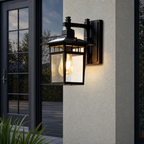 Black outdoor wall lantern with glass panels glowing on a light stucco exterior.