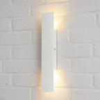 Slim white vertical wall light glowing warmly on a white brick wall.