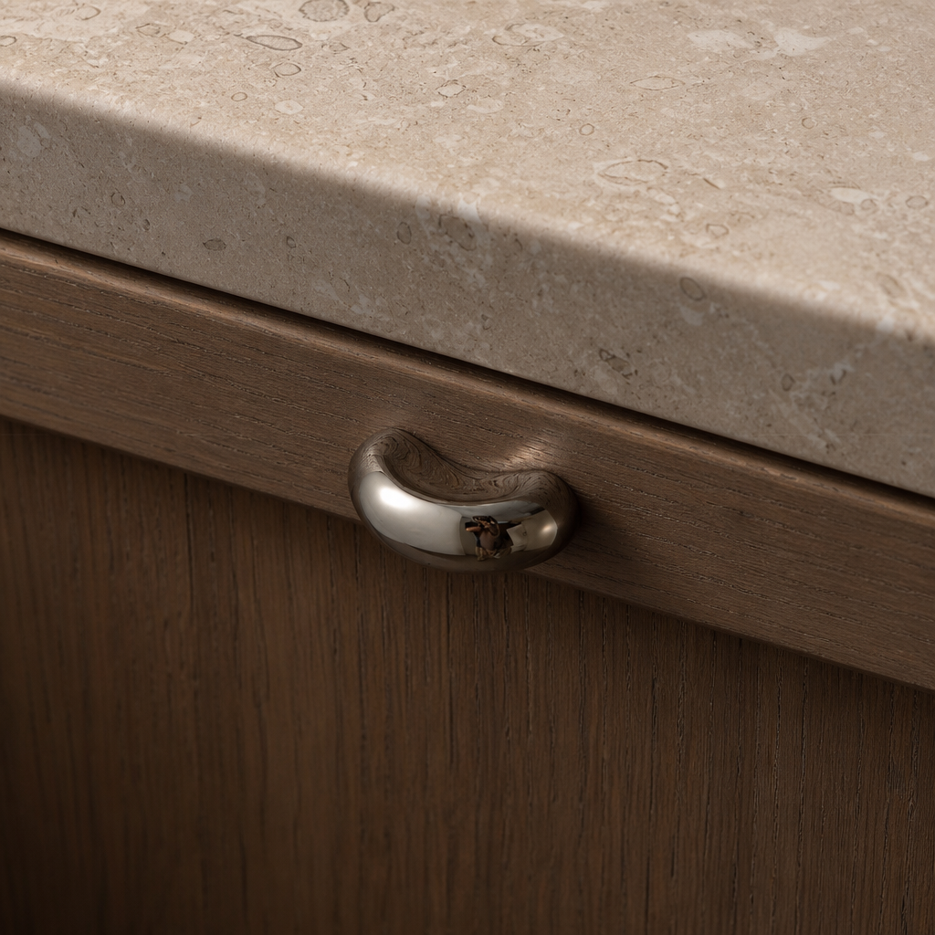 Polished oval metal drawer pull on a wood cabinet beneath a beige stone countertop.