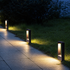 Three black rectangular pathway lights illuminate a tiled garden walkway at dusk.