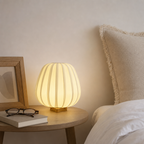 Warm ribbed table lamp glowing on a bedside table beside neutral bedding.