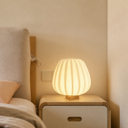 Glowing ribbed cream bedside lamp on a wooden nightstand beside a bed.