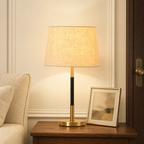 Lit table lamp with beige fabric shade, black and brass stem, and round brass base on a wooden nightstand.