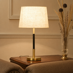 Lit table lamp with cream fabric shade, black and brass stem, and round brass base on a wooden side table.