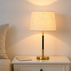 Lit table lamp with beige shade, black and gold stem, and round gold base on a white nightstand.