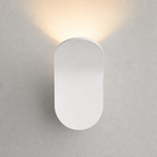 White capsule wall sconce emitting a warm upward glow on a pale wall.
