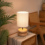 Glowing table lamp with a white fabric shade and rounded wooden base on a rattan cabinet.