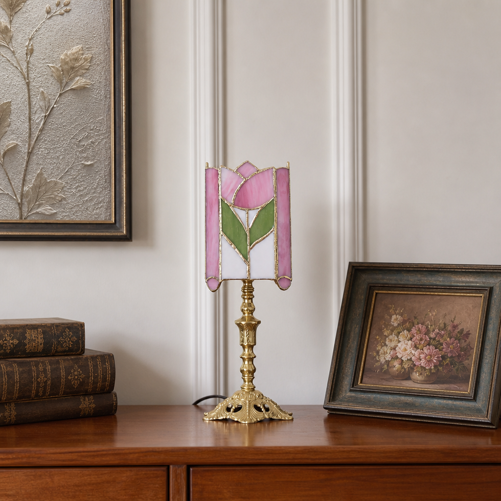 Pink rose stained-glass table lamp with ornate brass base on a wooden cabinet.