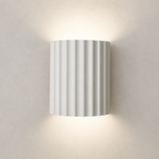 White ribbed cylindrical wall sconce glowing warmly on a plain wall