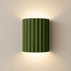 Olive green ribbed wall sconce glowing softly on a plain light wall.
