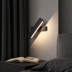 Black angled wall lamp glowing warmly above a bedside table with an open book.