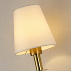 Warm lit brass wall lamp with a cream tapered shade on a neutral wall.