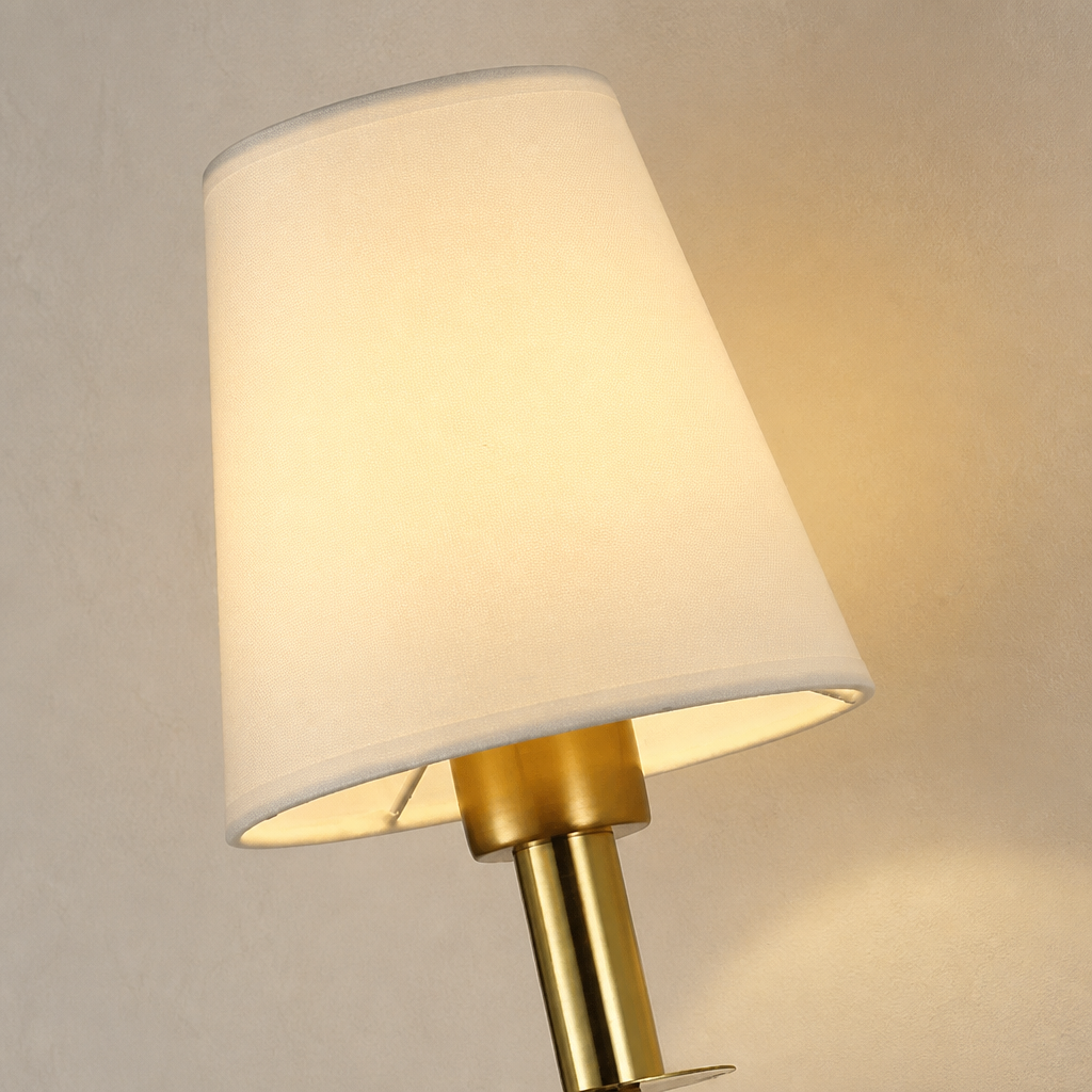 Warm lit brass wall lamp with a cream tapered shade on a neutral wall.