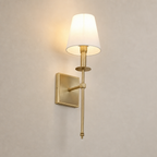 Brass wall sconce with a glowing cream shade on a plain light gray wall.