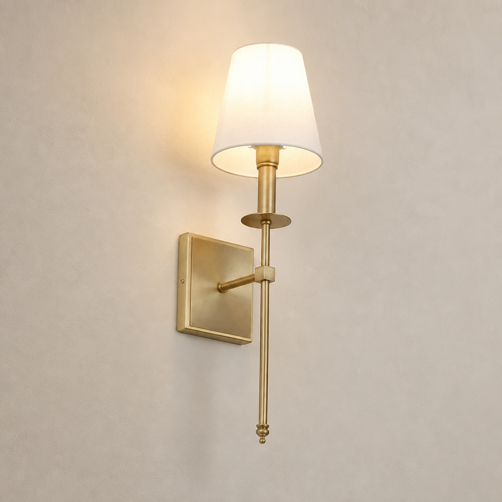 Brass wall sconce with a glowing cream shade on a plain light gray wall.