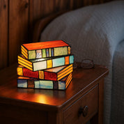 Stacked stained-glass book lamp glowing on a wooden bedside table.