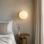 Glowing round wall lamp above a rustic wood bedside table beside a neutral bed.