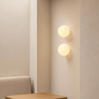 Two glowing round wall lights mounted vertically on a cream wall above a wooden bedside surface.