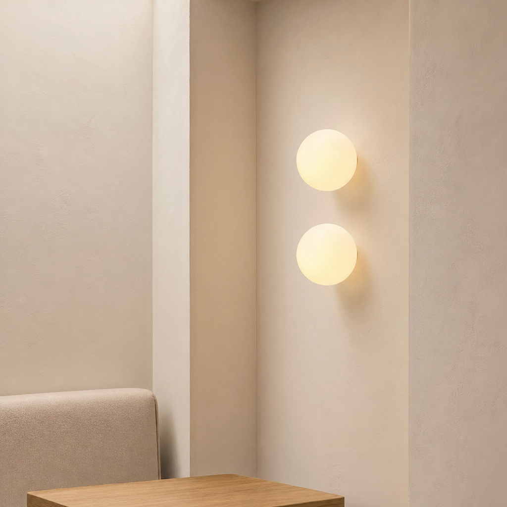 Two glowing round wall lights mounted vertically on a cream wall above a wooden bedside surface.
