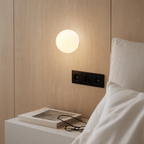 Glowing round wall lamp above a bedside table with a book and glasses next to white pillows.