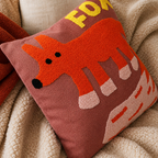 Dusty rose cushion with a tufted orange fox design on a cozy neutral blanket.