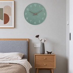 Sage green round wall clock with white numerals and copper hands above a wooden bedside table.