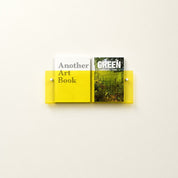 Yellow wall-mounted acrylic shelf holding two books on a pale wall.