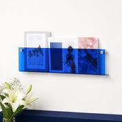 Blue transparent wall-mounted magazine holder with books and magazines on a white wall.