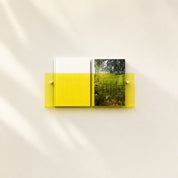 Yellow wall-mounted acrylic shelf holding two books on a pale minimalist wall.