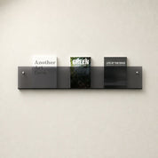 Smoked gray wall-mounted acrylic book shelf holding three magazines on a plain white wall.