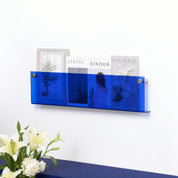 Blue wall-mounted acrylic magazine rack holding assorted magazines with flowers below.